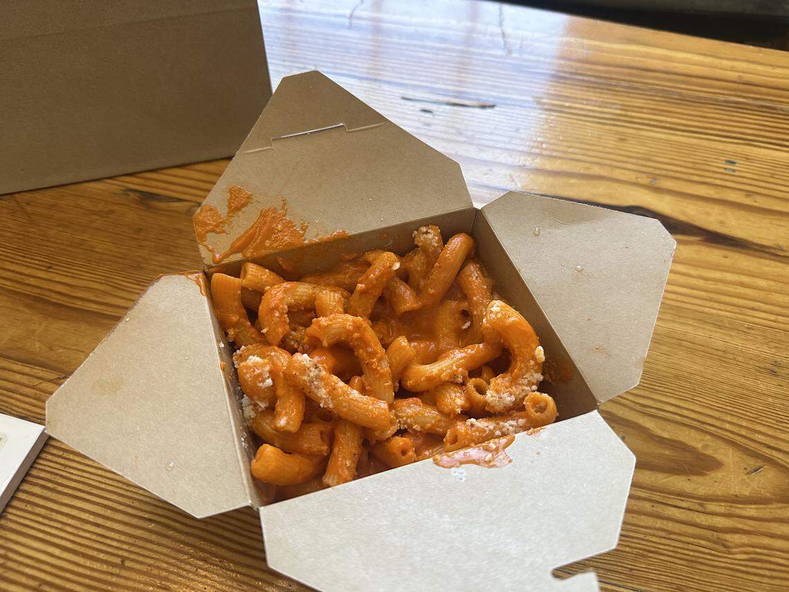 A high-angle shot of a cardboard takeout container sitting open on a light-colored wooden table. Inside the container is penne pasta thoroughly coated in a vibrant orange vodka sauce and lightly dusted with white parmesan cheese.